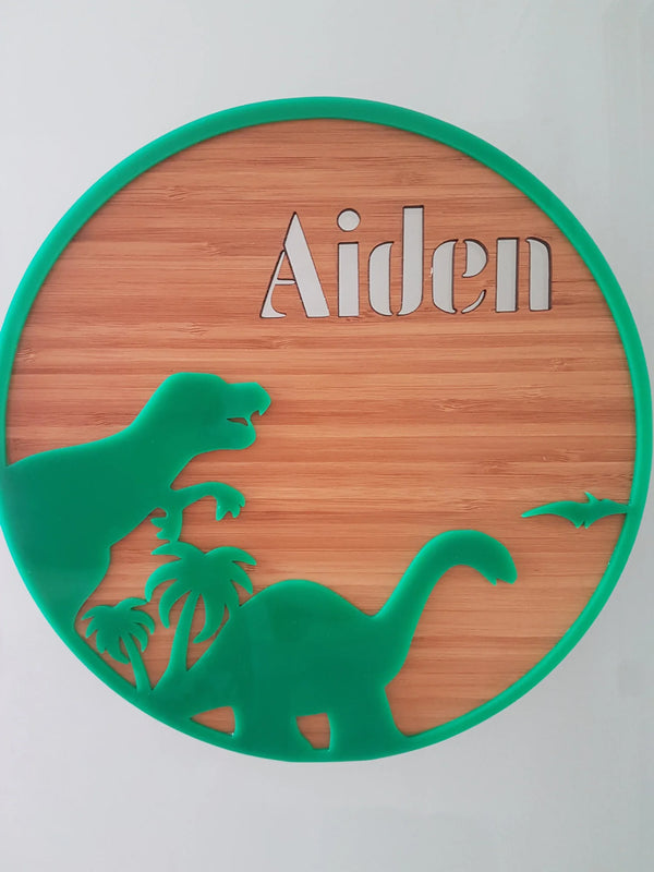 Round bamboo name plaque reading Aiden with green acrylic dinosaurs and palm trees