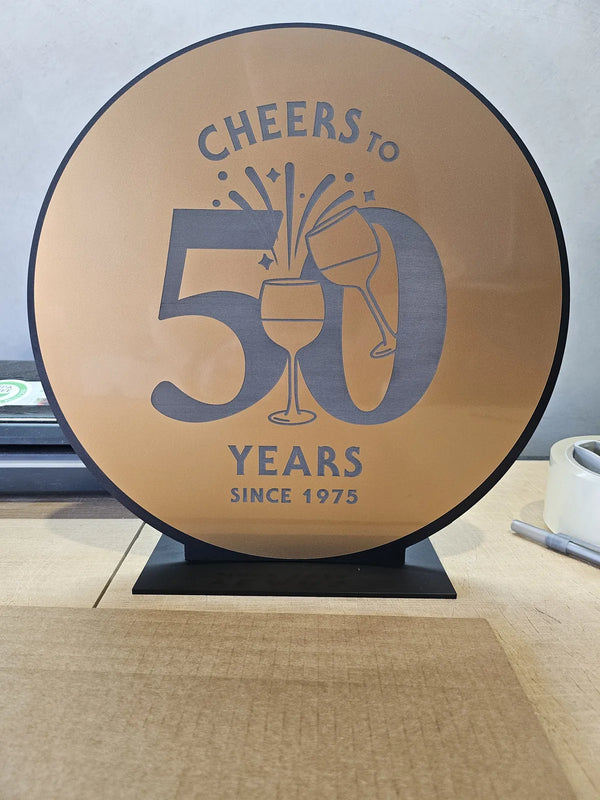Round gold and charcoal Cheers to 50 Years sign on a black stand