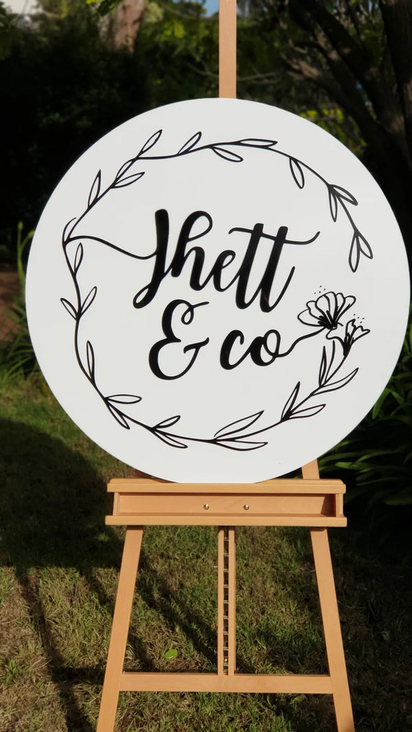 Round white acrylic wedding sign reading Jhett & co with a leaf wreath, on a wooden easel