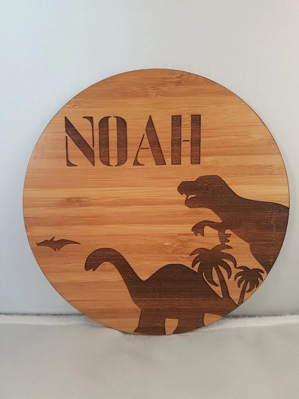 Round bamboo name plaque reading Noah with engraved dinosaurs and palm trees