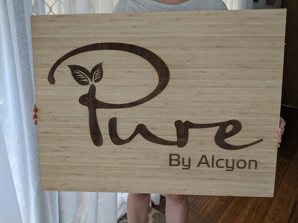 Large bamboo business sign engraved with the Pure by Alcyon logo, held by hand