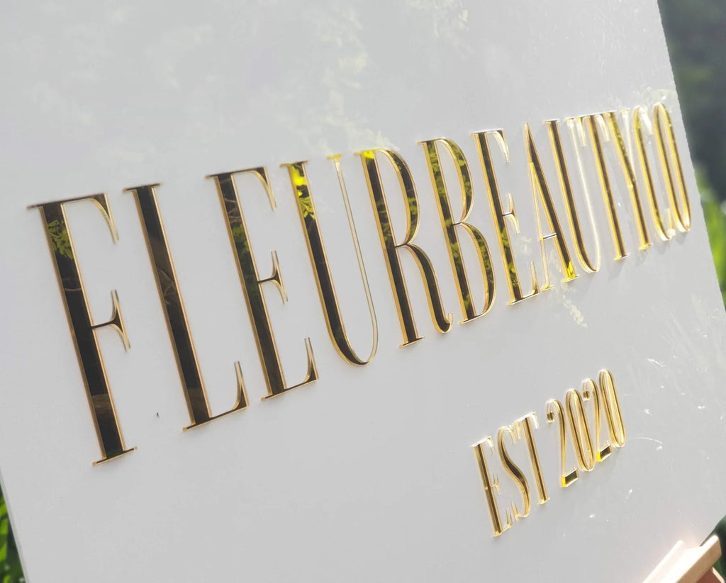 Close-up of gold mirror acrylic lettering on a white FLEURBEAUTYCO Est 2020 sign