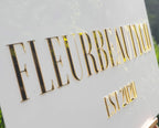Close-up of gold mirror acrylic lettering on a white FLEURBEAUTYCO Est 2020 sign