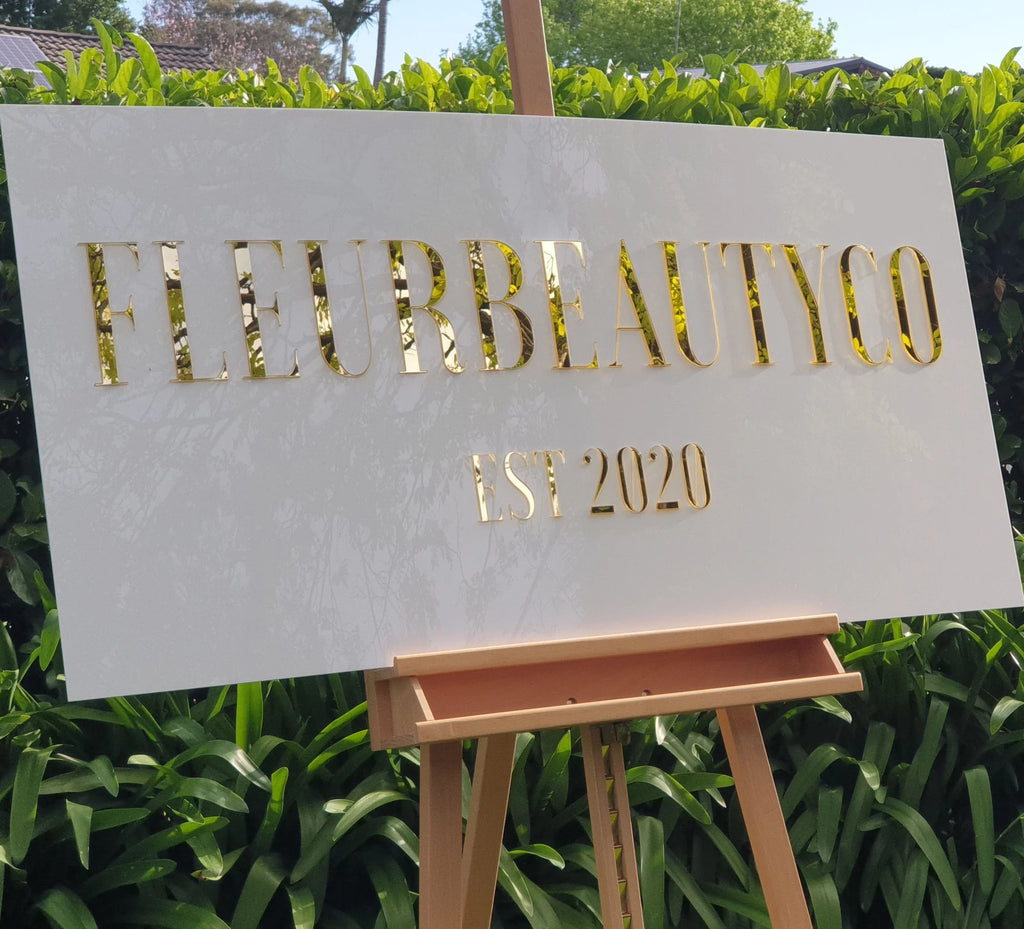 White rectangular FLEURBEAUTYCO Est 2020 sign with gold mirror lettering on a timber easel