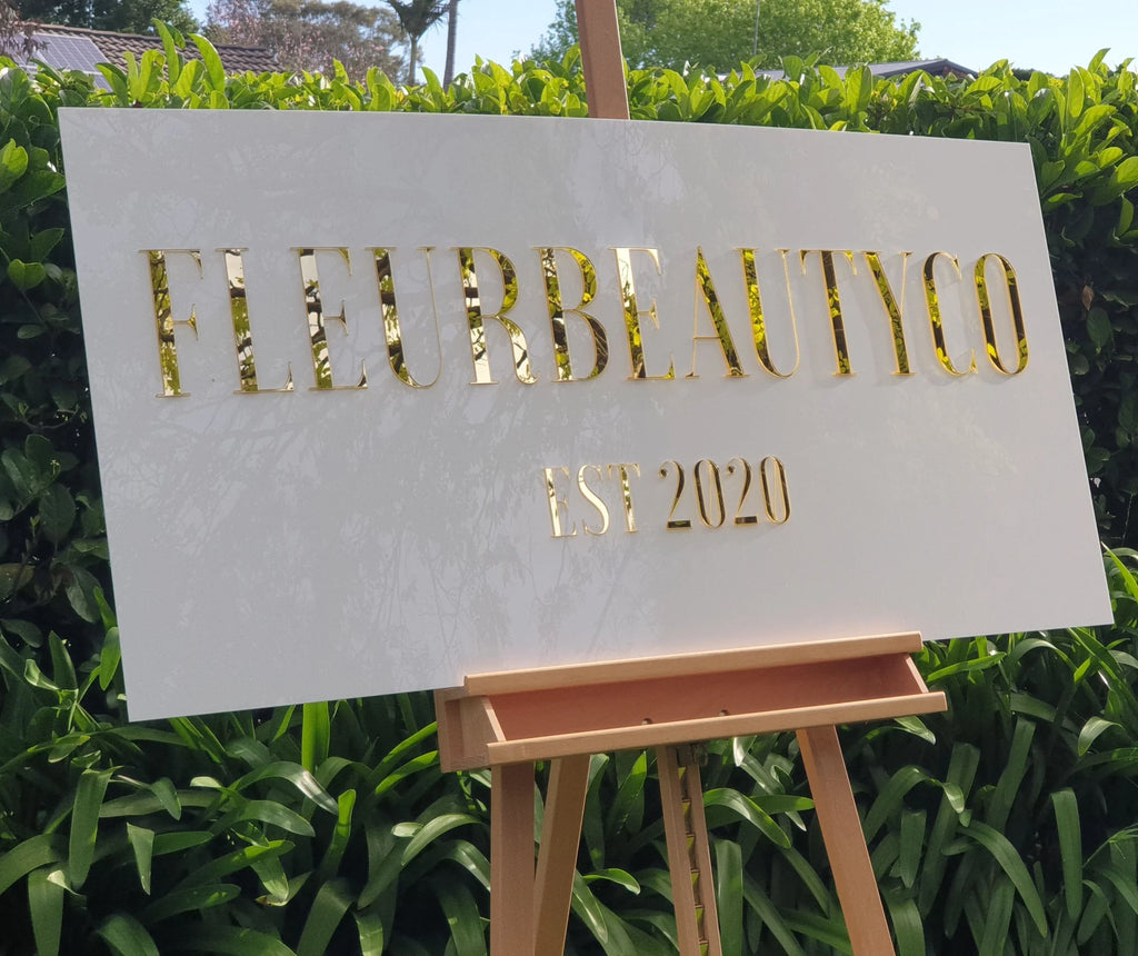 White rectangular FLEURBEAUTYCO Est 2020 sign with gold mirror lettering on an easel in front of a hedge