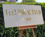 White rectangular FLEURBEAUTYCO Est 2020 sign with gold mirror lettering on an easel in front of a hedge