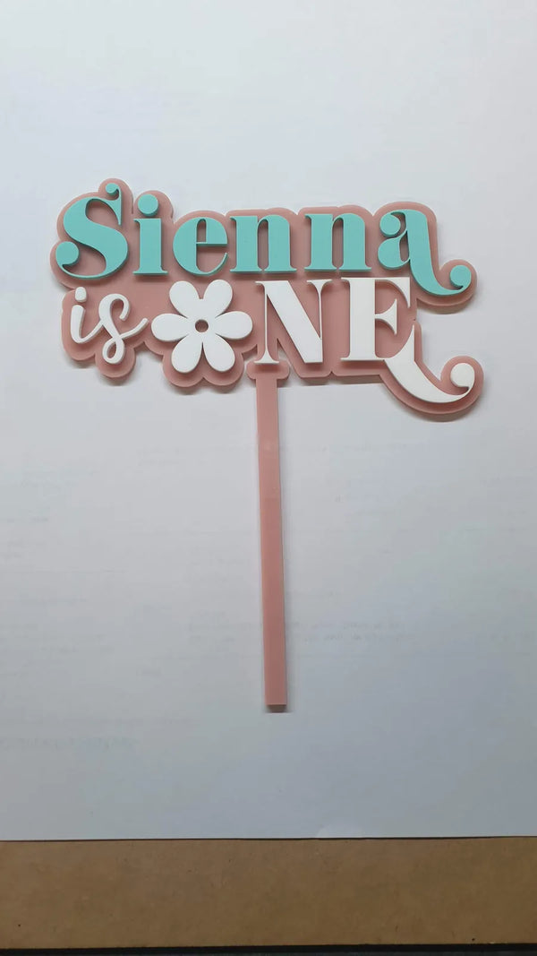 Teal and pink layered cake topper reading Sienna is ONE